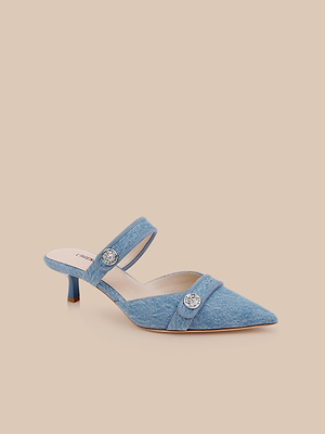 The Gilliane Denim Mule features light blue denim, a pointed toe, low heel, two straps with round silver decorative buttons, and a cushioned memory foam insole for comfort, shown on a beige background.