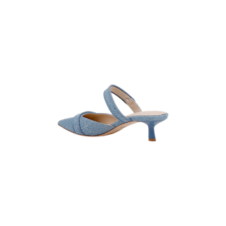 The Gilliane Denim Mule features light blue, textured denim with a pointed toe, crisscross front strap, and an angled mid-height heel. Designed for comfort with a memory foam insole, this stylish heeled mule is displayed against a plain white background.