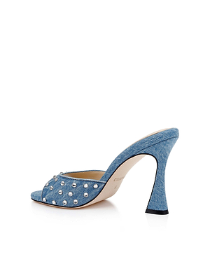 The Lucienne Embellished Denim Mule, a blue denim open-toe heel with silver studs, curved hourglass heel, and memory foam cushion for comfort, is shown against a white background.