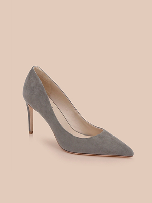The Maelle Suede Pump in smoke grey features a pointed toe and sleek stiletto heel, elegantly set against a light peach background.
