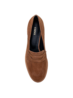 The Avriel Brogue Loafer, shown from above, features tan suede with brogueing and a penny slot detail. Its almond toe design is paired with a black insole displaying the L'AGENCE brand name in white lettering.