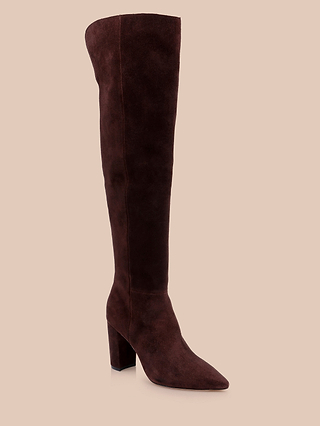 The Nanette Boot is a single over-the-knee boot crafted in mahogany brown suede, featuring a pointed toe and a chunky high heel, showcased against a beige background.
