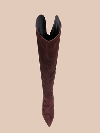 The Nanette Boot, a mahogany brown suede over-the-knee design, is viewed from above to highlight its pointed toe and black interior lining, set against a plain beige background.