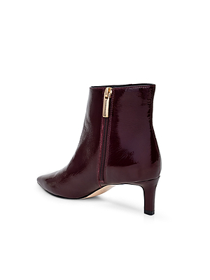 The L'AGENCE Lilly Kitten-Heel Bootie features glossy burgundy patent leather, a pointed toe, gold side zipper, and a low kitten heel, shown against a white background.