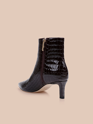 The Lilly Kitten-Heel Bootie, a black city boot crafted from crocodile-embossed leather with a side zipper and a mid-height heel, is shown against a beige background.