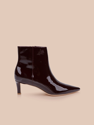 La bottine Lilly à talon « kitten heel » est une bottine de ville en cuir verni brillant bordeaux foncé, dotée dun bout pointu et dun talon « kitten heel » court et fin, présentée sur un fond beige.