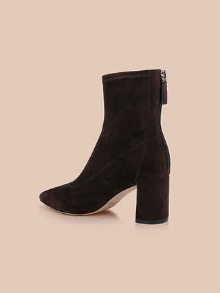 The Salome Stretch Suede Boot, a dark brown ankle boot featuring a chunky block heel and a back zipper, is displayed on a beige background.