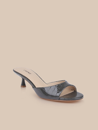 The Arleen Kitten-Heel Mule is a black open-toe, slip-on heeled sandal crafted from crocodile embossed leather with a textured pattern and a low kitten heel. It features a memory foam insole for added comfort and is displayed on a light beige background.
