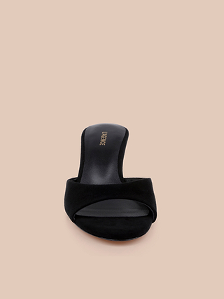 The Arleen Kitten-Heel Mule is a black suede sandal featuring a single wide strap and a low kitten heel, displayed from the front against a beige background—an elegant and comfortable women’s shoe ideal for any occasion.