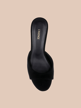The Arleen Kitten-Heel Mule is a black suede open-toe slide sandal featuring a single wide strap and a black insole with the LAGENCE brand name in gold lettering, displayed on a beige background. This comfortable women’s shoe offers effortless style with its chic kitten heel design.