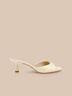 The Arleen Kitten-Heel Mule is a cream-colored, open-toe sandal featuring a low, curved kitten heel, shown against a beige background.