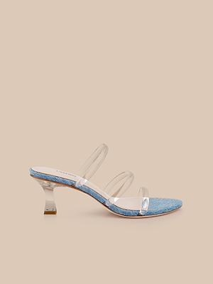 The Ludovica Kitten-Heel Sandal features a light blue textured footbed, three clear top straps, and a transparent curved heel, shown against a neutral beige background.