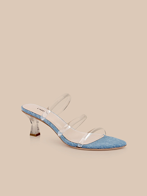 The Ludovica Kitten-Heel Sandal features a pointed toe, three clear vinyl straps, and a petite hourglass heel, embodying minimalist style against a beige background.
