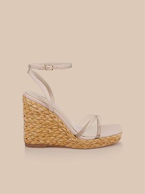 The Malone Wedge Espadrille features a braided raffia wedge heel, clear toe straps, and an ankle strap with buckle, shown on a light beige background.
