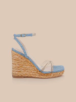 The Malone Wedge Espadrille features a woven straw heel, light blue denim details, a transparent toe strap, and a matching blue ankle strap with buckle, shown against a plain beige background.