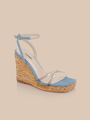 The Malone Wedge Espadrille features a woven raffia wedge sole, light blue denim accents on the toe and heel, a clear plastic toe strap, and a light blue buckled ankle strap, shown against a beige background.
