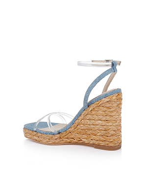 The Malone Wedge Espadrille features a denim upper, woven straw heel, transparent ankle strap, and clear toe straps, shown against a white background.