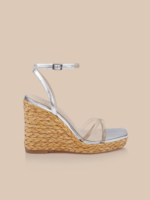 The Malone Wedge Espadrille, featuring a braided raffia heel, clear toe straps, and a metallic leather ankle strap with buckle, is shown against a plain beige background.
