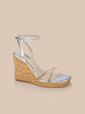 The Malone Wedge Espadrille features a silver ankle strap, clear toe strap, metallic leather sole, and braided raffia wedge heel, shown on a beige background.