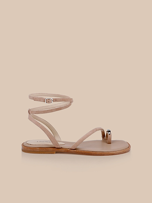 The Mariana Suede Sandal features tan suede, asymmetric straps, a nude toe loop with a silver accent, delicate ankle ties, and a flat wooden sole, beautifully displayed on a beige background.