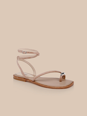 The Mariana Suede Sandal features a minimalist tan suede design with asymmetric straps, a thin buckled ankle strap, and a silver-accented toe loop, shown against a light beige background.