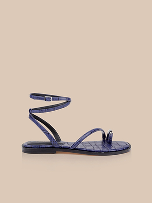 The Mariana Leather Sandal features stylish purple crocodile-textured leather, an ankle strap, toe loop, and a metallic accent on the toe strap—ideal for those who love the navy croc-embossed sandal trend. Displayed on a beige background.