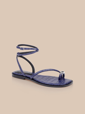 The Mariana Leather Sandal features a purple faux-crocodile finish with a thin ankle strap and toe loop, inspired by ankle tie flats, shown against a plain beige background.