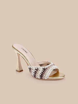 The Moyes Crochet Sandal showcases a chic woven crisscross strap design in cream, brown, and gold, with a sculpted gold metallic heel and sole. Presented on a beige background for an elegant look.