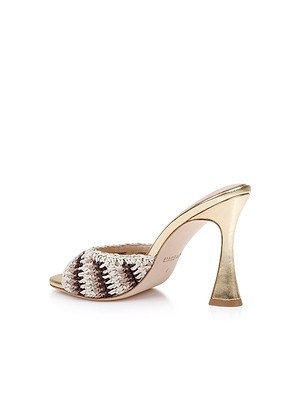 The Moyes Crochet Sandal is a gold mule with high heels, featuring a cream and brown woven open-toe upper, a sculpted shiny metallic heel, and a beige sole. Photographed on a white background.
