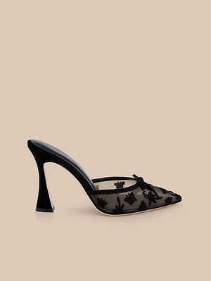The Prasad Mesh Mule is a black mesh mule with a pointed toe, floral and butterfly embroidery, and a bow detail, shown on a beige background.