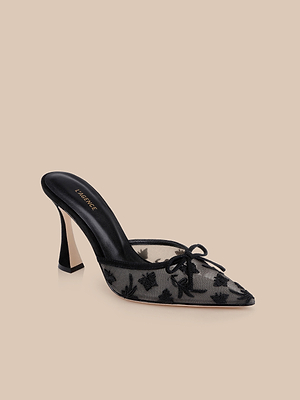 The Prasad Mesh Mule features a black mesh design with a pointed toe, decorative bow, butterfly embroidery, black heel, and Versace logo on the insole, shown against a beige background.