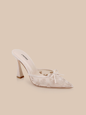 The Prasad Mesh Mule features a beige pointed toe, high heel, translucent mesh upper with delicate butterfly embroidery and a small bow. A curved heel and matching beige background complete this elegant, stylish design.