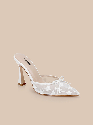 The Prasad Mesh Mule is a white, pointed-toe shoe featuring a sheer mesh upper, delicate butterfly embroidery, and a small bow on top, shown against a neutral beige background.