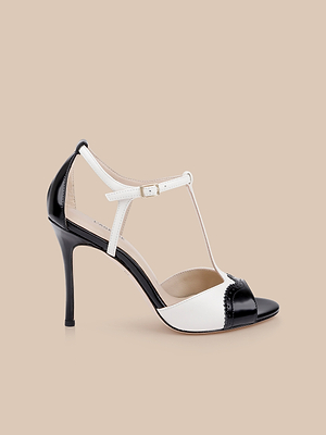 The Radha Leather Sandal is a black and white high-heeled shoe featuring a T-strap design, brogue details, peep toe, and buckle closure, shown on a beige background.