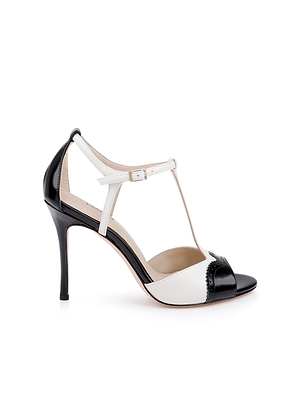 The Radha Leather Sandal is a high-heeled, black and white T-strap sandal with an ankle buckle, open toe, and classic brogue detailing on the toe and heel.