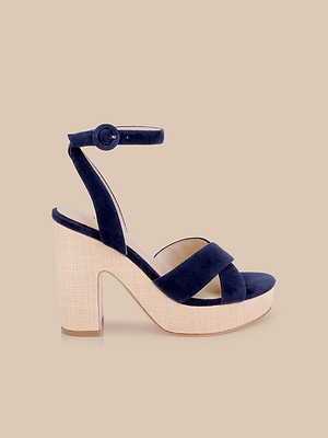 The Sandrine Platform Sandal features navy blue velvet crisscross straps, an ankle strap with buckle, and a chunky raffia-wrapped wedge heel and platform, shown on a beige background.