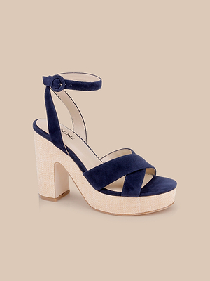 The Sandrine Platform Sandal features navy suede with crisscross straps, an ankle buckle, and a beige textured sole. Its block heel and raffia-wrapped wedge details evoke modern elegance and warm-weather style, shown on a light beige background.