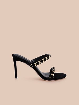 The Sarika Chain Sandal is a black suede high-heeled sandal featuring two straps with gold chain accents, showcased on a plain beige background.