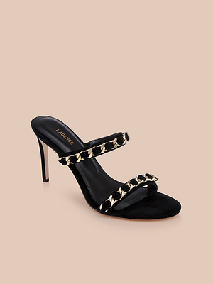 The Sarika Chain Sandal is a black suede high-fashion heel with two straps, each featuring bold gold chain accents, shown against a plain beige background.