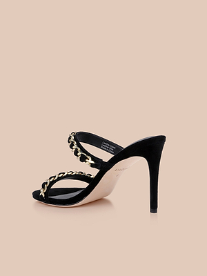 Displayed on a beige background, the Sarika Chain Sandal features black suede, two straps embellished with gold chain hardware, and a high-fashion heel.