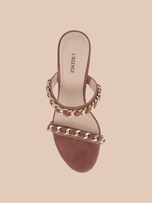 The Sarika Chain Sandal is a single mauve suede heel with two straps adorned with gold chain hardware, displayed from above on a light beige background.