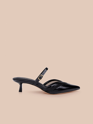 The Shelbie Kitten-Heel Mule, a black leather pointed-toe style with a thin ankle strap, buckle, and cutout side detail, is displayed on a plain beige background.