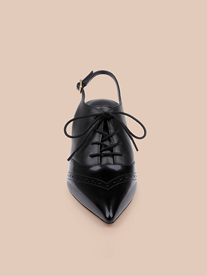 The Shreya Leather Slingback Pump is a black leather stiletto with a pointed toe, lace-up front, decorative stitching, and an ankle strap with buckle, shown from above on a beige background.