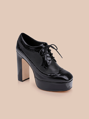 The Tania Oxford Pump, a black patent leather shoe featuring brogue details, a chunky block heel, rounded toe, and lace-up front, is displayed against a plain beige background.