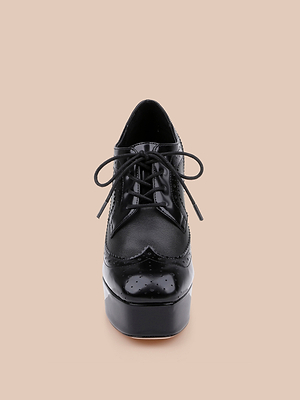 The Tania Oxford Pump is a shiny black platform shoe with laces and a bold block heel, shown from the front on a plain beige background.