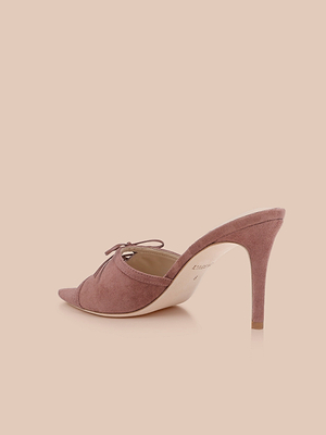 The Thames Suede Sandal, a mauve high-heeled mule featuring pointed toe, side cutout, and bow detail, is displayed on a light peach background, combining elegance with the sophistication of a stiletto heel.