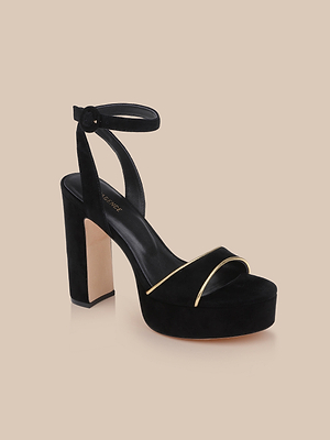 The Essie Platform Sandal is a black suede shoe with a thick heel, ankle strap, and gold accent on the toe strap, showcased against a beige background.