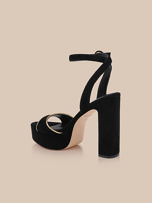 The Essie Platform Sandal, a black suede shoe with a thick block heel, ankle strap, open toe, and gold circular accent near the front, is displayed on a beige background.