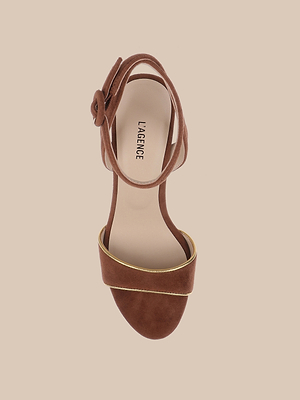 The Essie Platform Sandal features brown suede, a wide front strap with gold piping trim, an ankle strap, and a beige insole with the L’AGENCE logo. Its displayed on a light beige background.