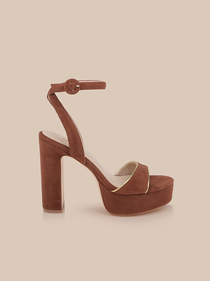 The Essie Platform Sandal is a brown suede heeled sandal with an ankle strap, round buckle, platform sole, thick heel, and gold piping trim, set against a solid light beige background.